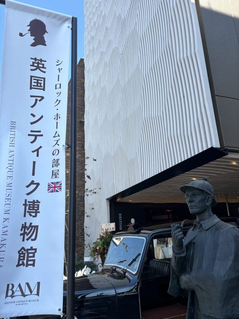 BAM鎌倉の外観。白い波打つ外壁の下に本物のロンドンタクシー（ブラックキャブ）が停まり、手前にパイプをくわえたシャーロック・ホームズのブロンズ像、左に英国アンティーク博物館の幟が立つ