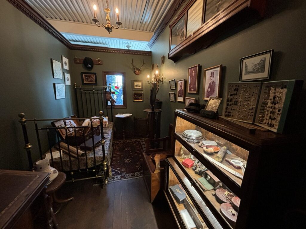BAM鎌倉のシャーロック・ホームズの部屋にある寝室。真鍮の寝台に開いたままの旅行トランクが置かれ、壁には肖像写真と鹿の角、ガラス扉の書棚に懐中時計やパイプが並ぶ