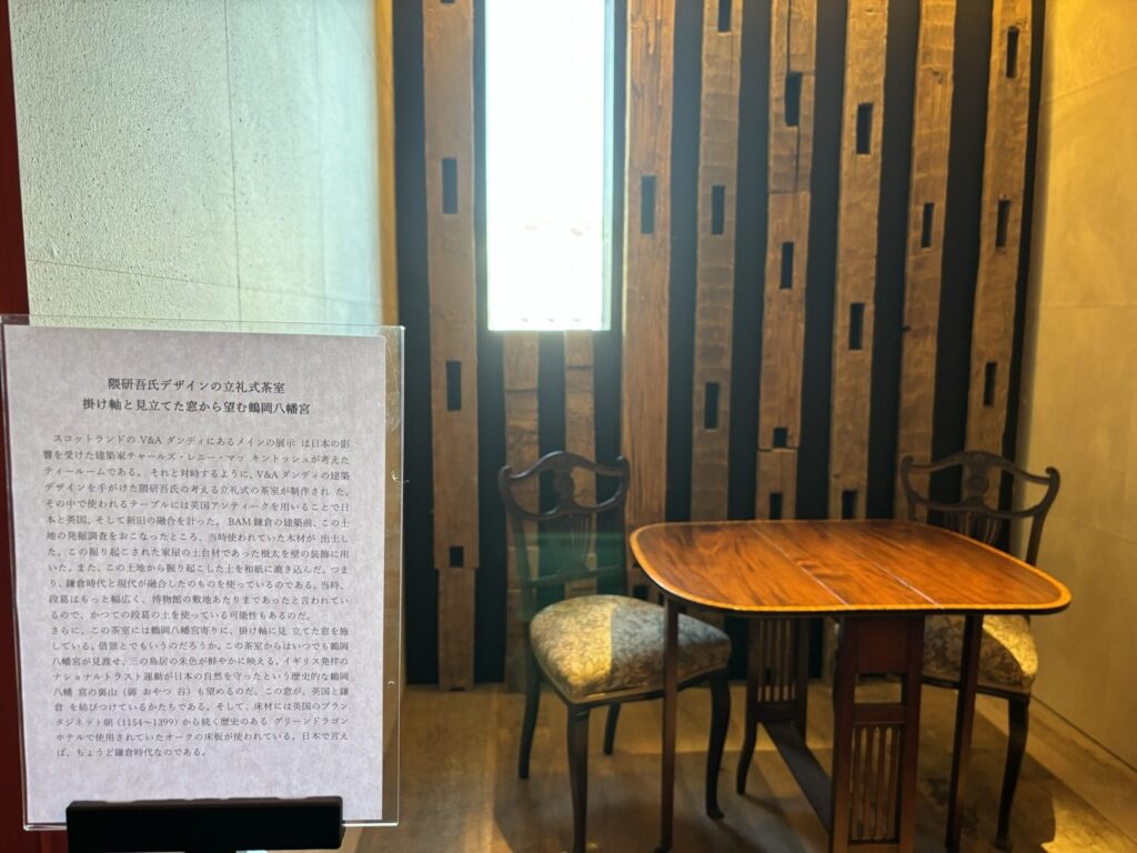 BAM鎌倉4階の隈研吾デザインの立礼式茶室。縦に並ぶ木材の壁と、掛け軸に見立てた細長い窓、英国アンティークのテーブルと椅子。手前に日本語の解説パネル