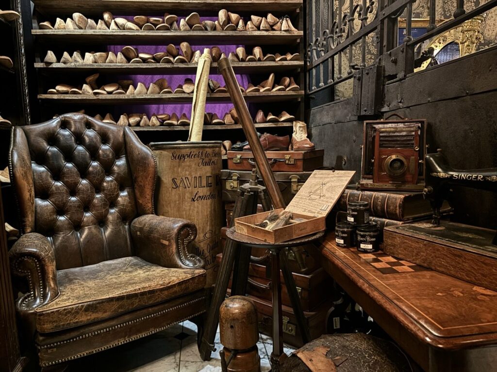 BAM鎌倉の展示。棚一面に並ぶ靴の木型と革のチェスターフィールドチェア、サヴィル・ロウと書かれた布袋、蛇腹の木製カメラ、ジョンロブの靴クリーム缶が置かれている