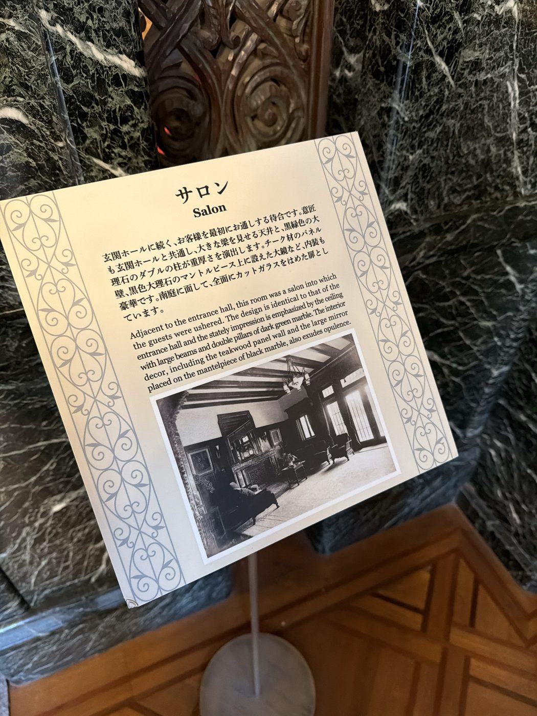 サロンの解説カード。玄関ホールに続く待合で、大きな梁を見せる天井、黒緑色の大理石のダブルの柱、チーク材のパネル壁、黒色大理石のマントルピース上の大鏡について説明されている